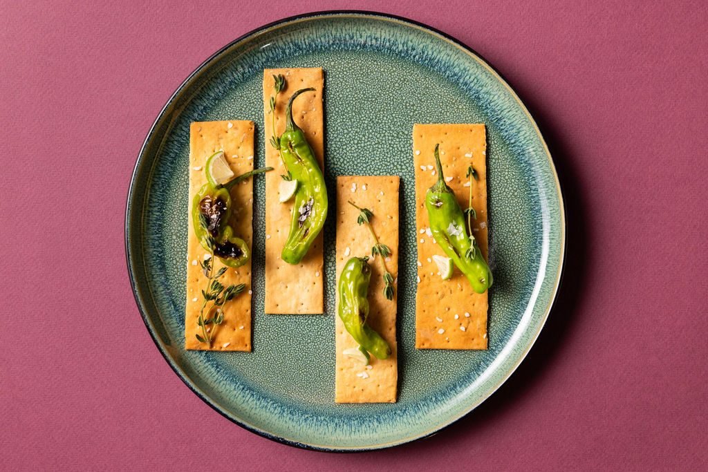 Blistered shishito peppers on Firehook crackers on a teal plate against a magenta backdrop