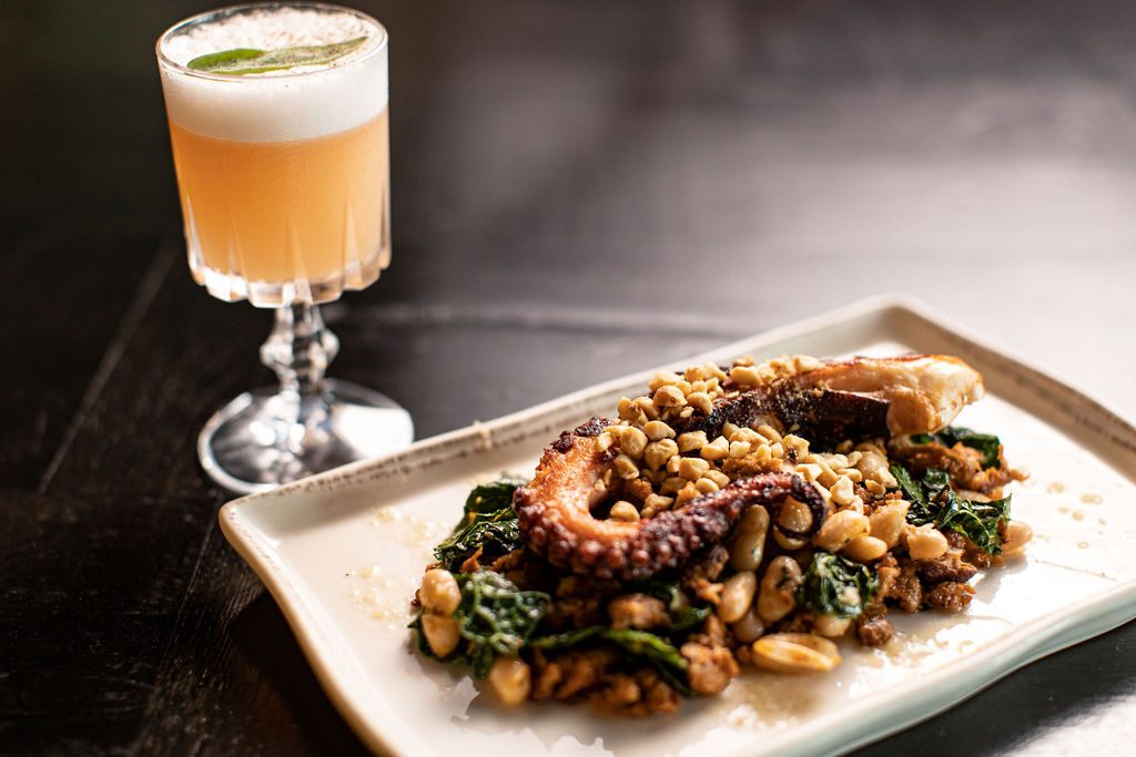 Grilled octopus over beans and greens on a stone plate with a sour cocktail behind