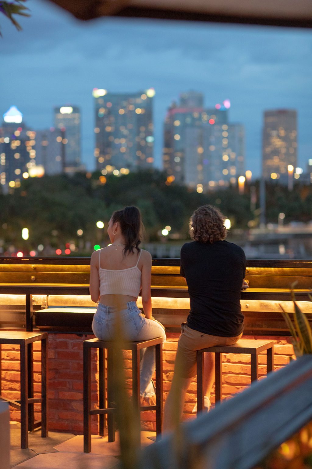 Couple on bar stools at a rooftop bar at dusk, the Tampa skyline lit up behind them