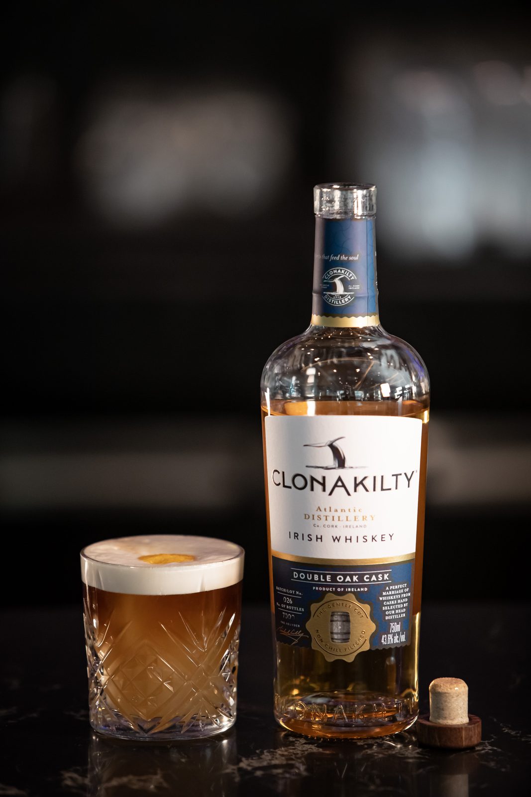 Bottle of Clonakilty Double Oak Cask Irish whiskey beside a whiskey sour with a foamed top on a dark marble bar